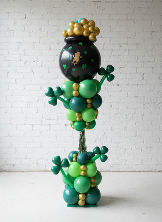 A decorative balloon display resembling a pot of gold with green and gold balloons, shamrocks, and a leprechaun hat on top.