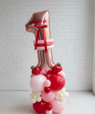 A balloon pedestal featuring a variety of colorful balloons with fruit designs, such as strawberries and blueberries, predominantly in red, white, and green shades, tied with red ribbons.