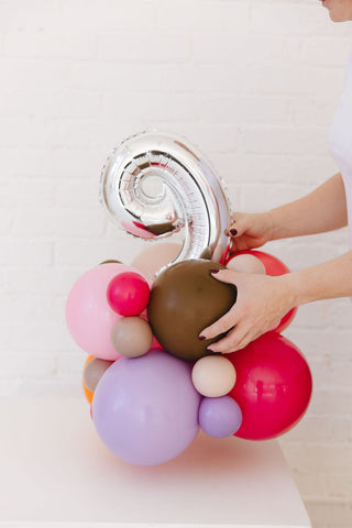 Colorful balloon arrangement with a hand holding a silver number balloon against a white background