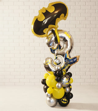 A balloon column featuring a superhero theme with a large bat emblem, yellow, silver, and black balloons, and decorative elements.