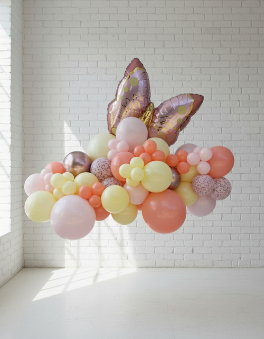 A colorful balloon garland featuring a variety of colors including pink, gold, and yellow, shaped into a semi-circular form with an Easter egg and butterfly design, displayed against a white wall.
