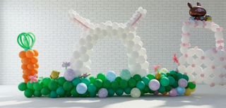 An Easter-themed backdrop featuring a bunny face, an Easter basket, and a carrot patch made from balloons, with a colorful array of eggs and a stuffed rabbit.